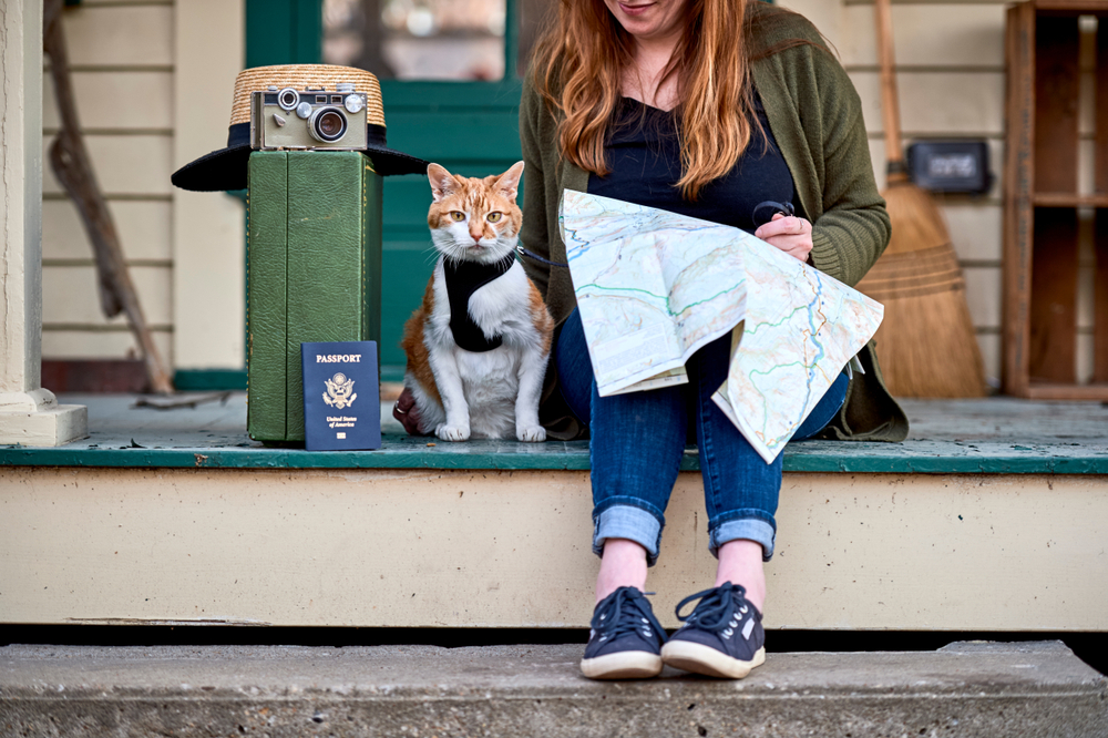 A woman sits on a porch holding a map, ready for her trip. Her cat, fresh from the vet and wearing a harness, sits contently beside her. Nearby are a suitcase, a hat with a camera on it, and a passport, evidence of their shared adventure about to unfold.