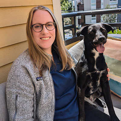 A person with long hair and glasses is sitting on a couch on a balcony, smiling. They have an arm around a black and white dog. The dog is sitting upright with its tongue out. There is a wooden wall and a view of buildings in the background.